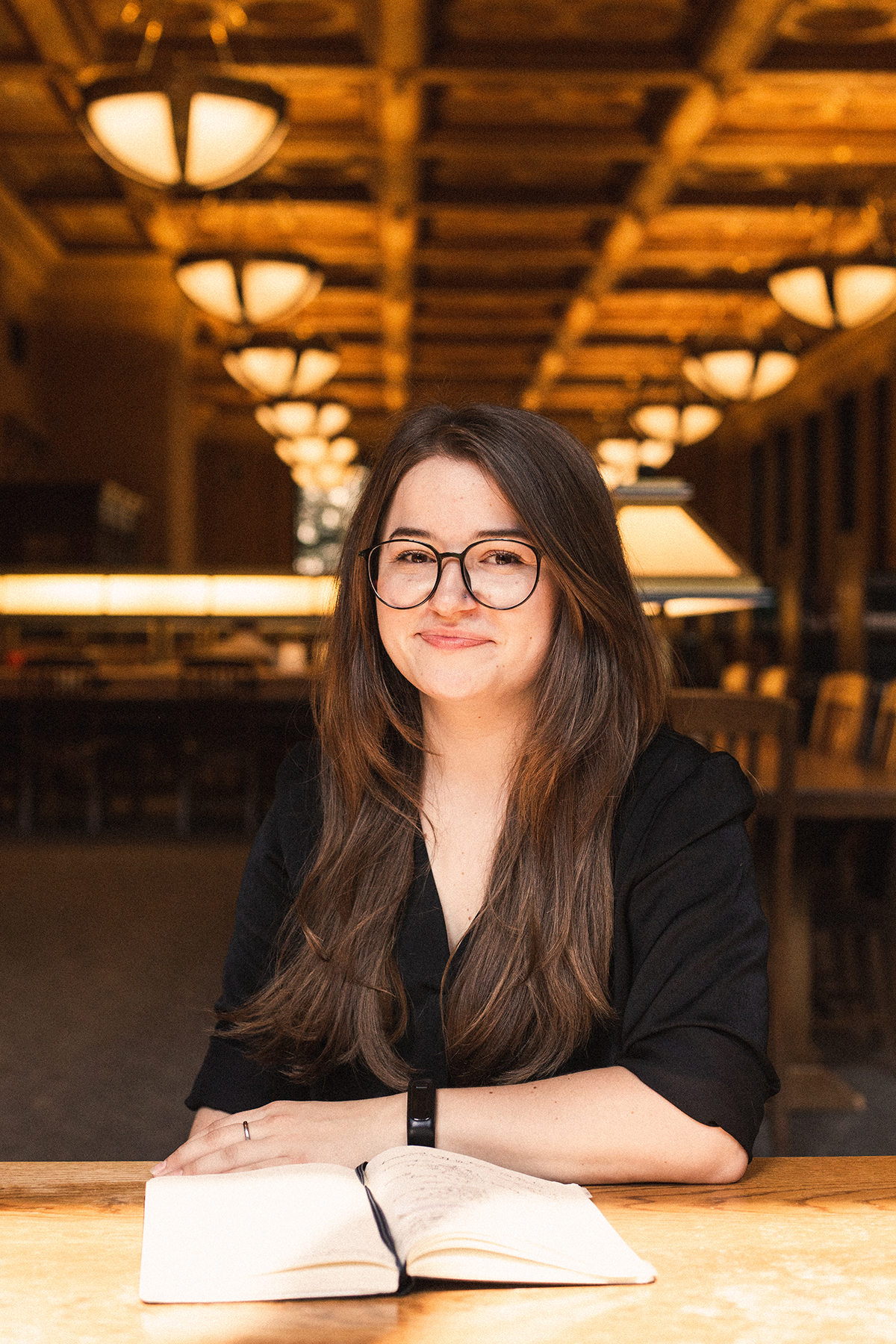 Portrait of a young woman with brown eyes, long brown hair, and large glasses wearing a black blouse. Sitting at a table in a library with high, ornate ceilings and warm lighting.